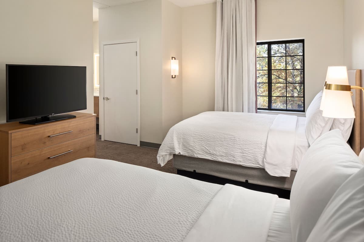 A hotel room with a flat-screen TV on a wooden dresser, two beds with white bedding, a window with a view of trees, and a wall-mounted lamp.