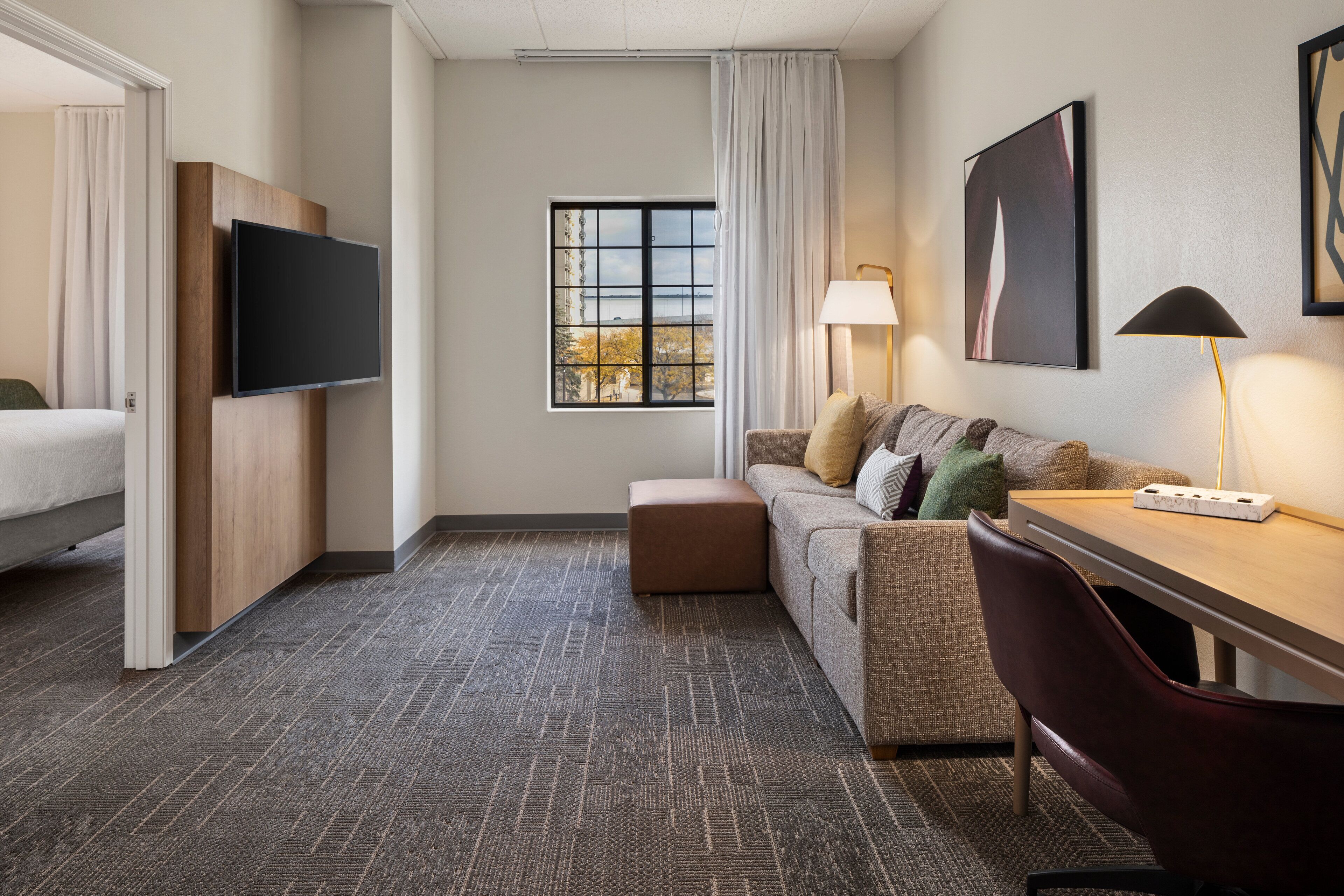Quarto de hotel com cama, sofá, escrivaninha, televisão, janela com vista e luminária.