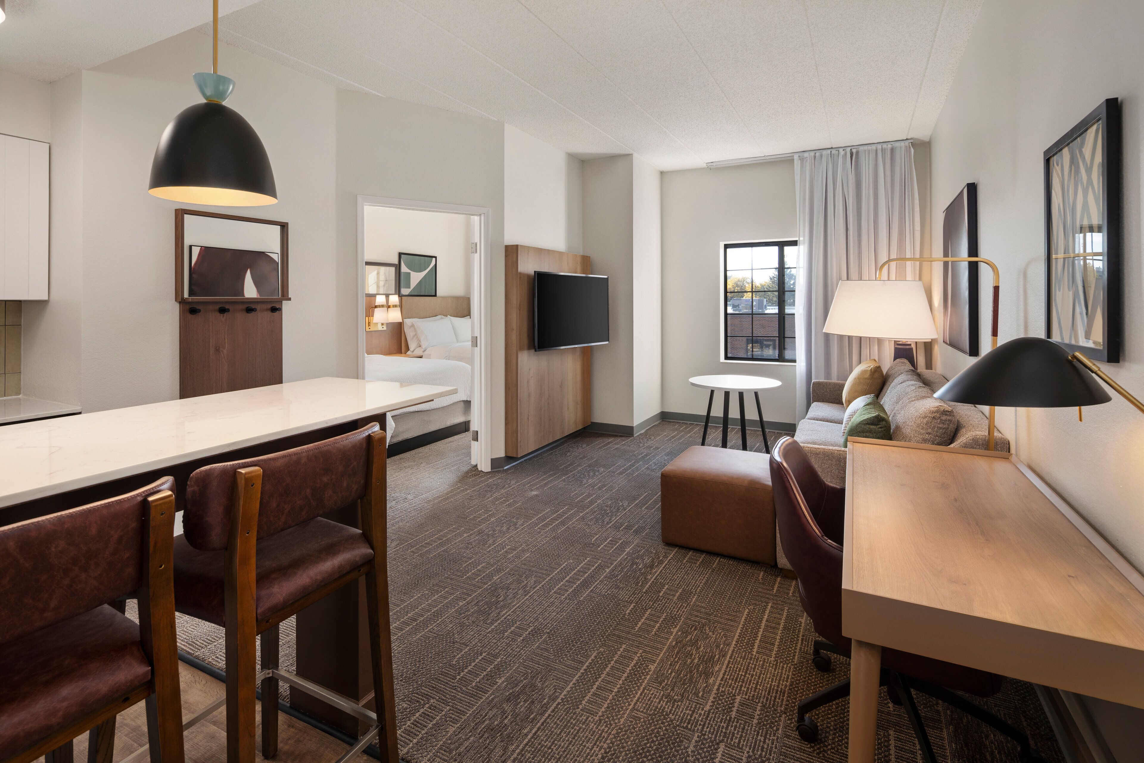 A hotel room with a kitchenette, a dining area with bar stools, a seating area with a sofa and armchairs, a TV mounted on the wall, and a window with curtains.