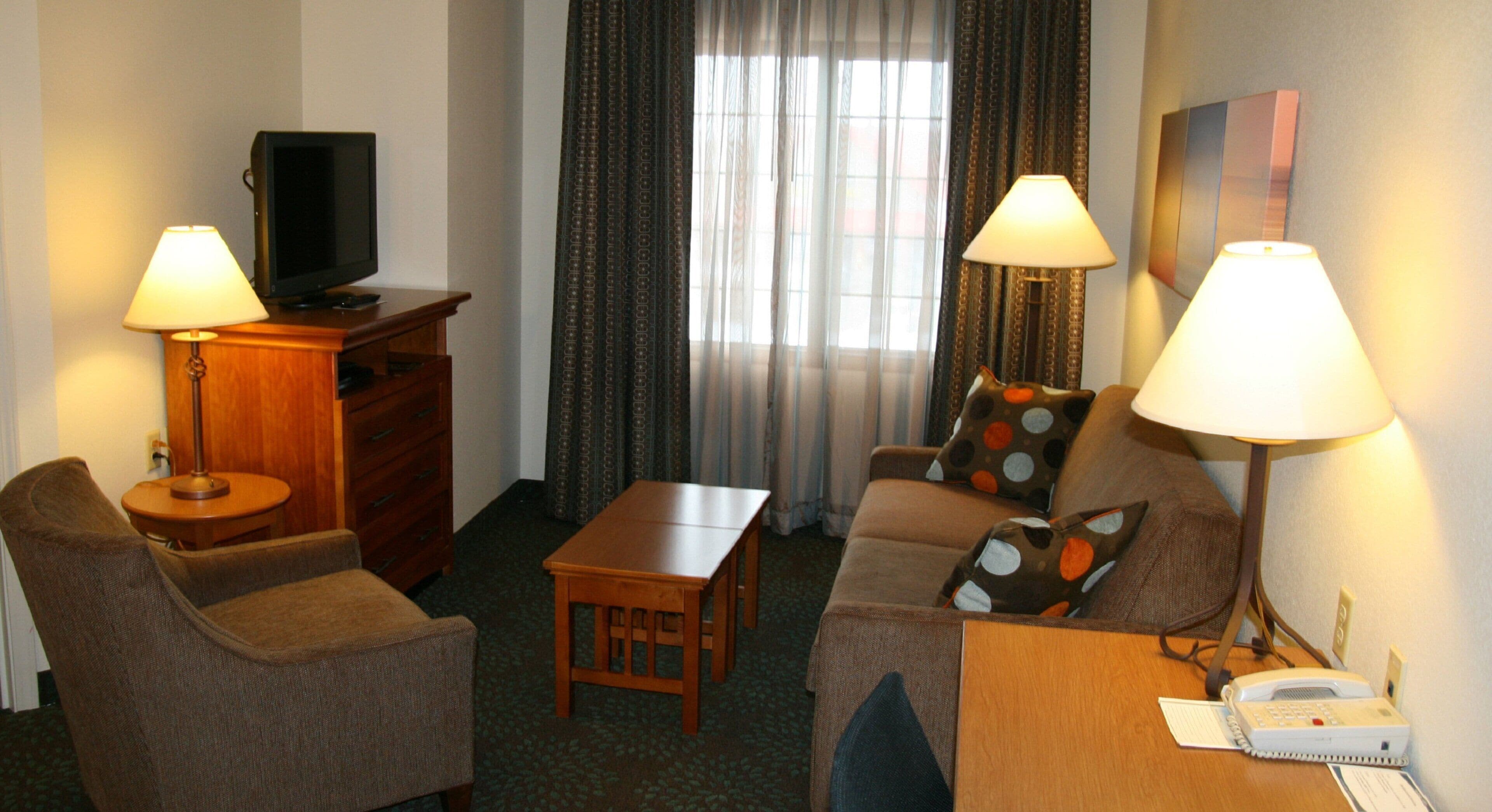 Quarto de hotel com sofá, duas poltronas, mesa de madeira pequena, televisão sobre aparador de madeira e duas luminárias.
