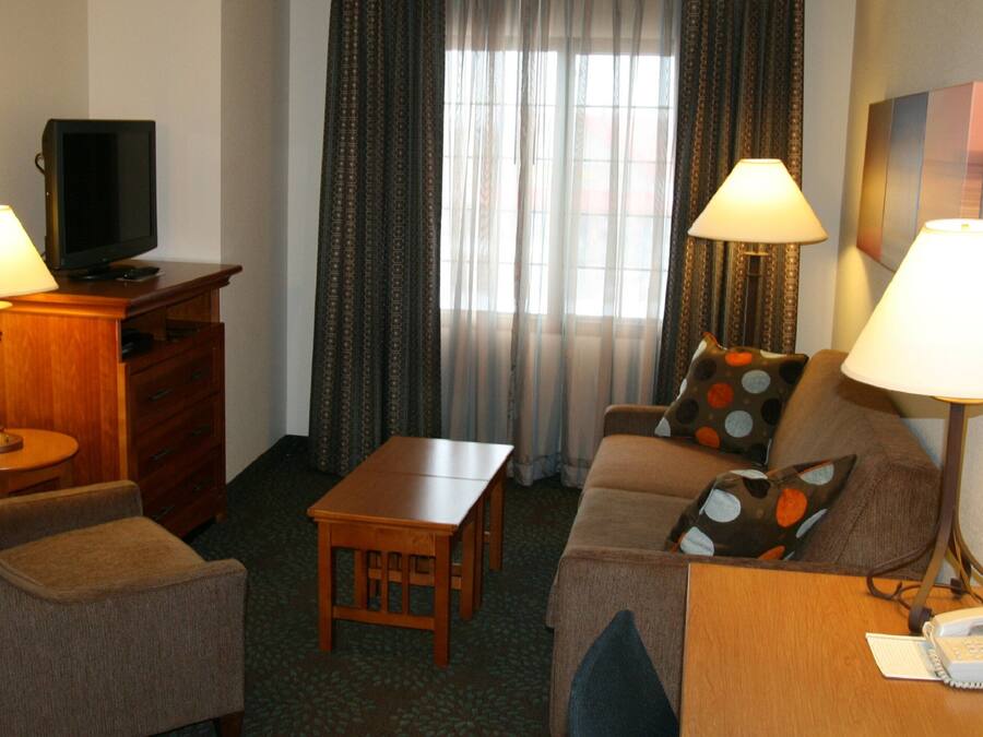 Quarto de hotel com sofá, duas poltronas, mesa de madeira pequena, televisão sobre aparador de madeira e duas luminárias.