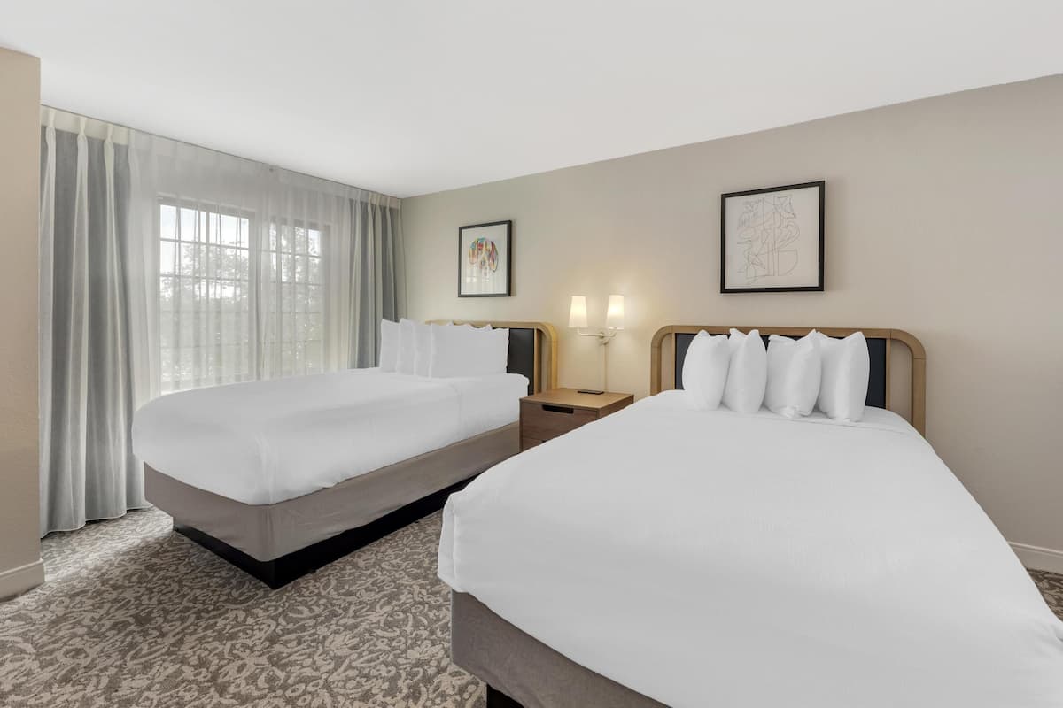 A hotel room with two beds, a large window with curtains, two wall-mounted lamps, and two framed pictures.