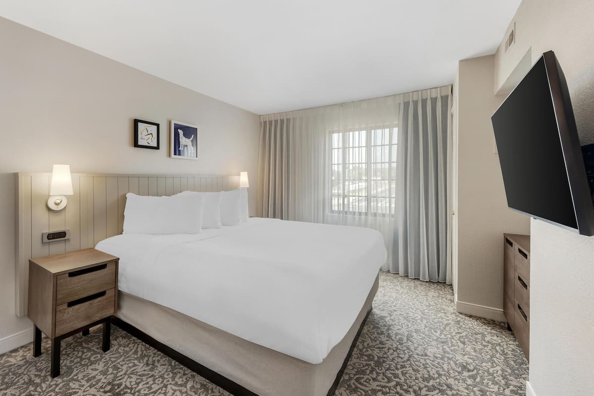 A hotel room with a large bed, bedside tables, a flat-screen TV, and a window with curtains.