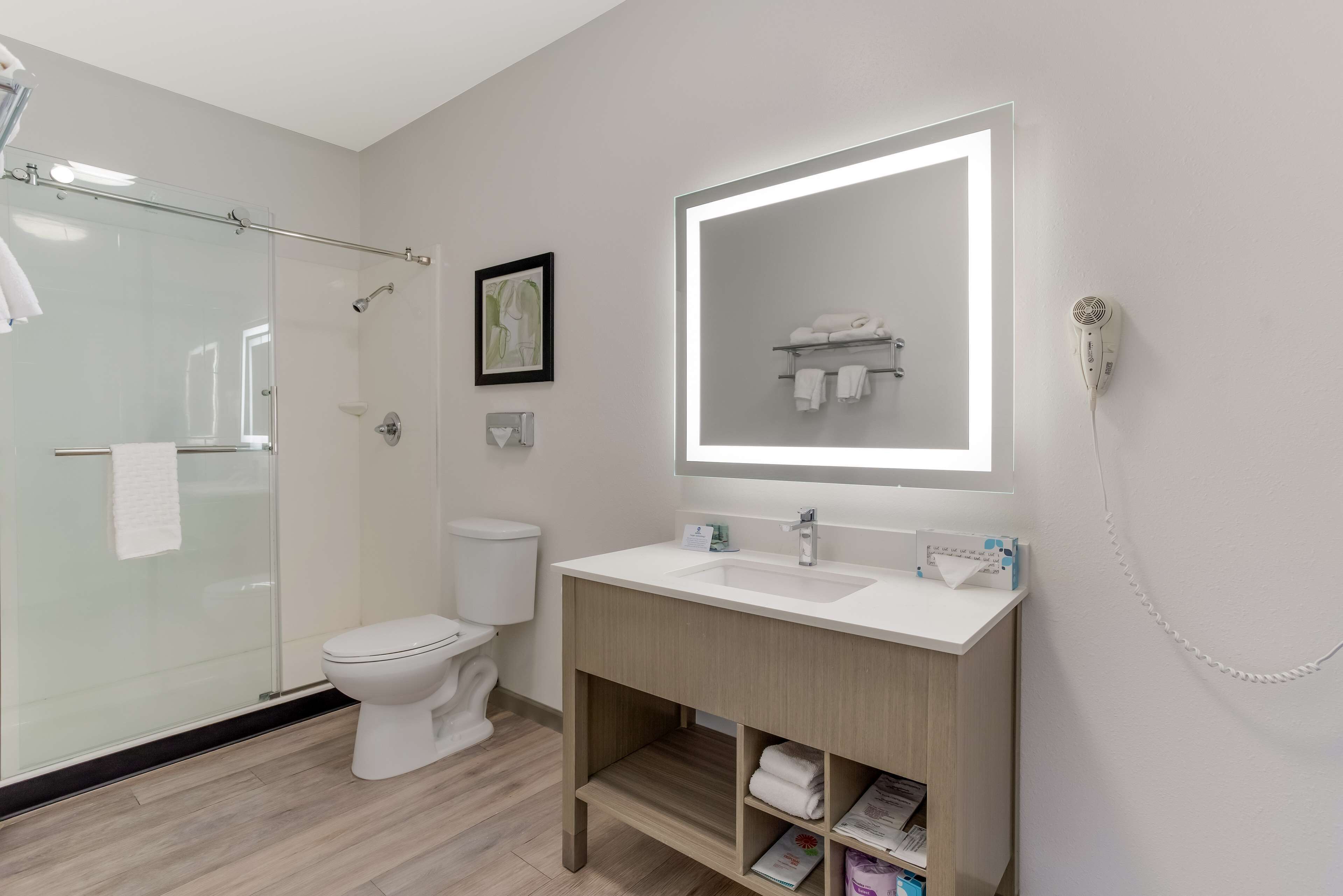 Une salle de bain moderne dotée d’une douche à parois de verre, de toilettes blanches, d’un meuble-lavabo avec un grand miroir et d’un sèche-cheveux fixé au mur.