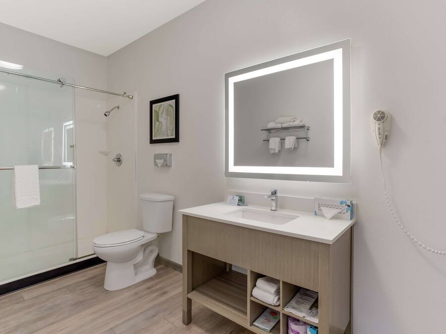 Une salle de bain moderne dotée d’une douche à parois de verre, de toilettes blanches, d’un meuble-lavabo avec un grand miroir et d’un sèche-cheveux fixé au mur.