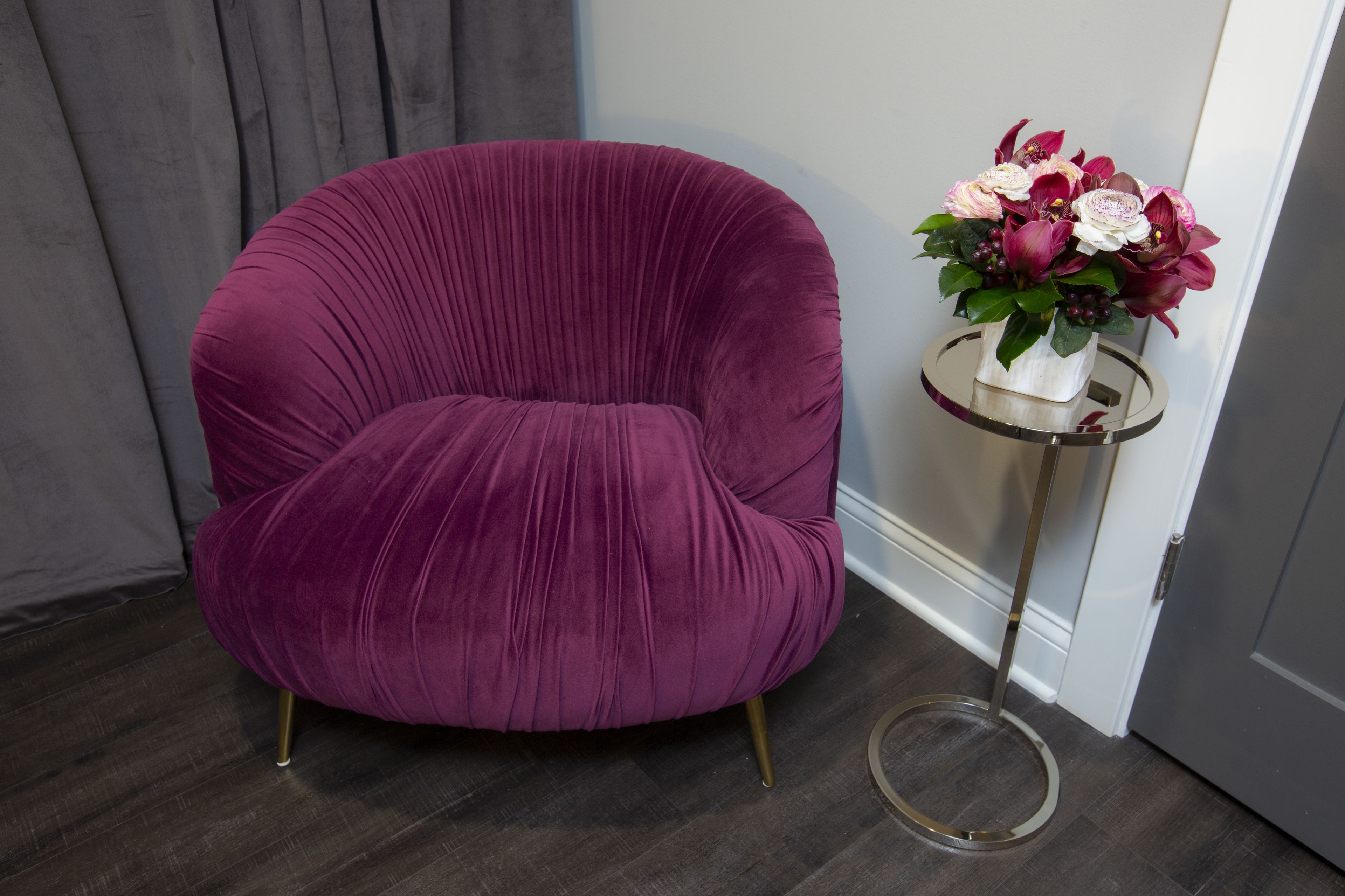 A plush, deep purple armchair with a round back and gold legs, positioned next to a small, round side table with a clear glass top. On the table, there is a white vase containing a bouquet of flowers with pink and white blooms.