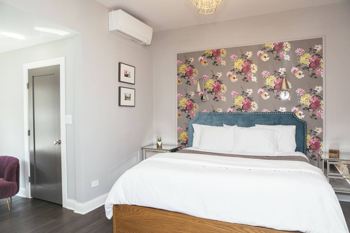 A bedroom with a bed, a nightstand, a chair, a floral-patterned wall, and a door.