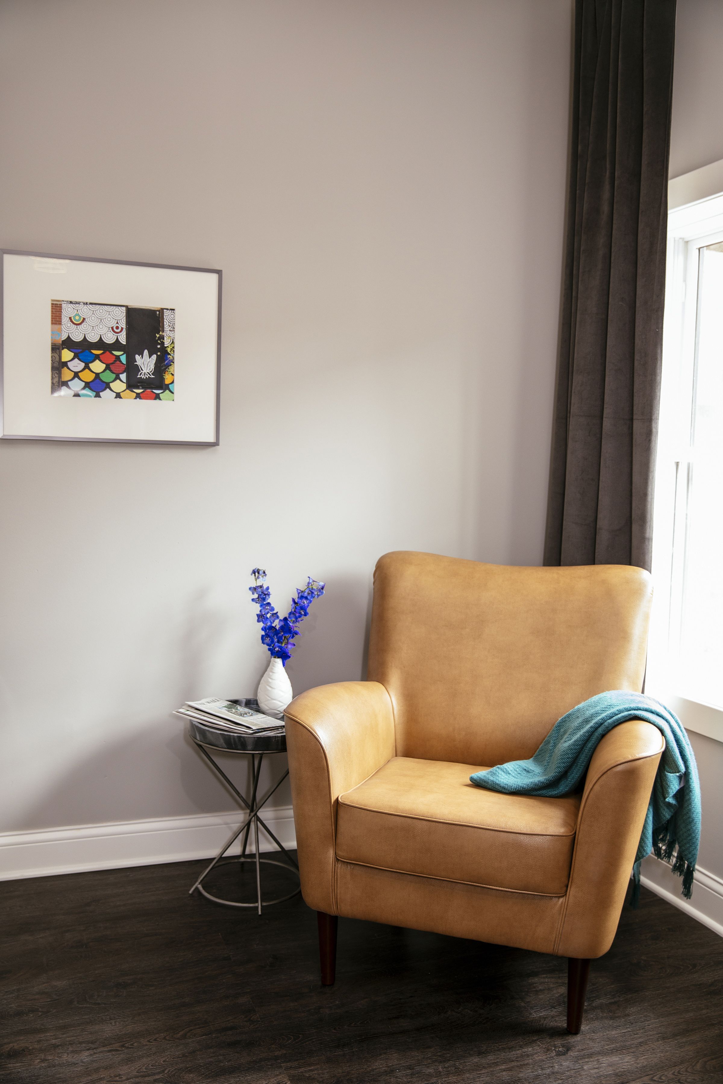 Un sillón de cuero color bronce con una manta azul aguamarina, un jarrón blanco con flores azules y una obra de arte abstracto enmarcada en la pared.