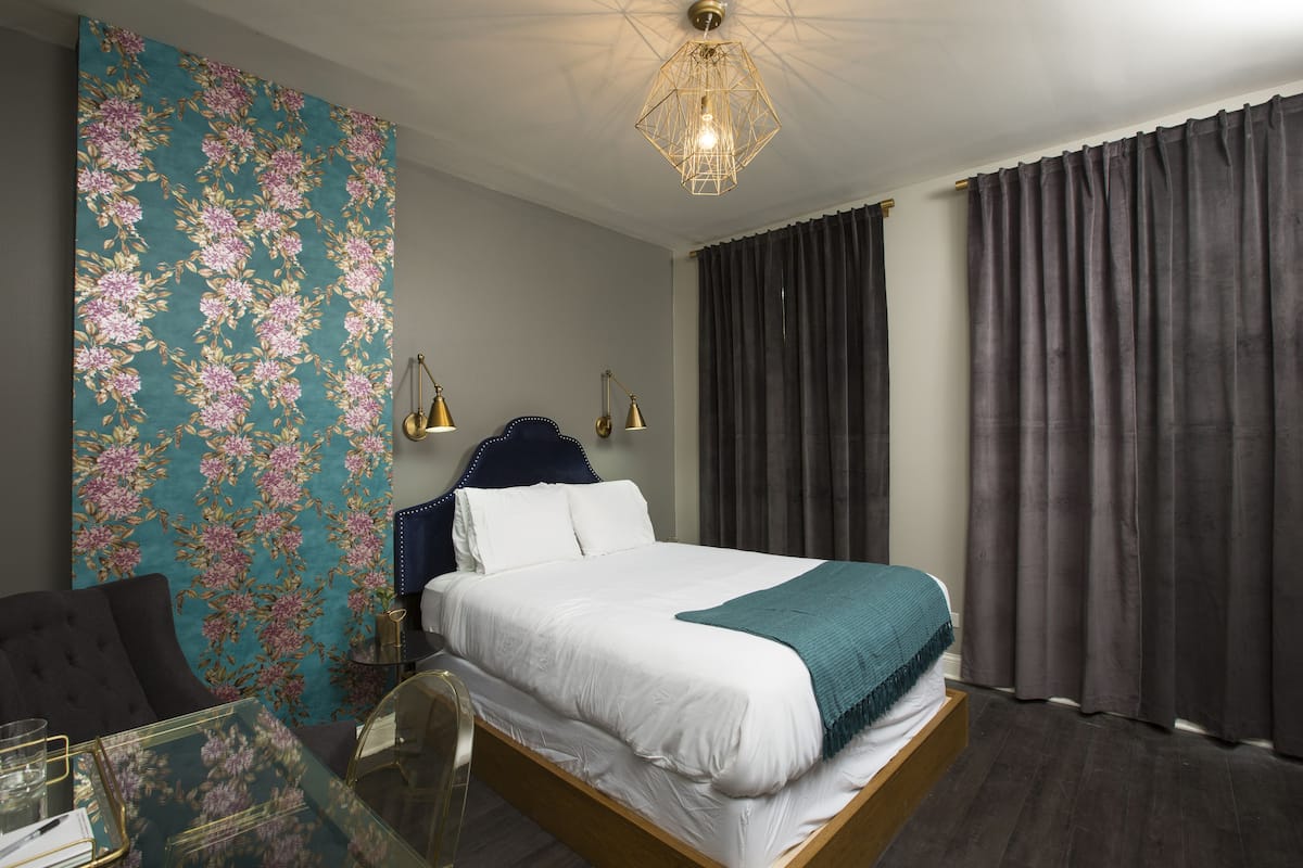 A hotel room with a bed, a glass table, a chair, a nightstand, and a wall with a floral pattern.