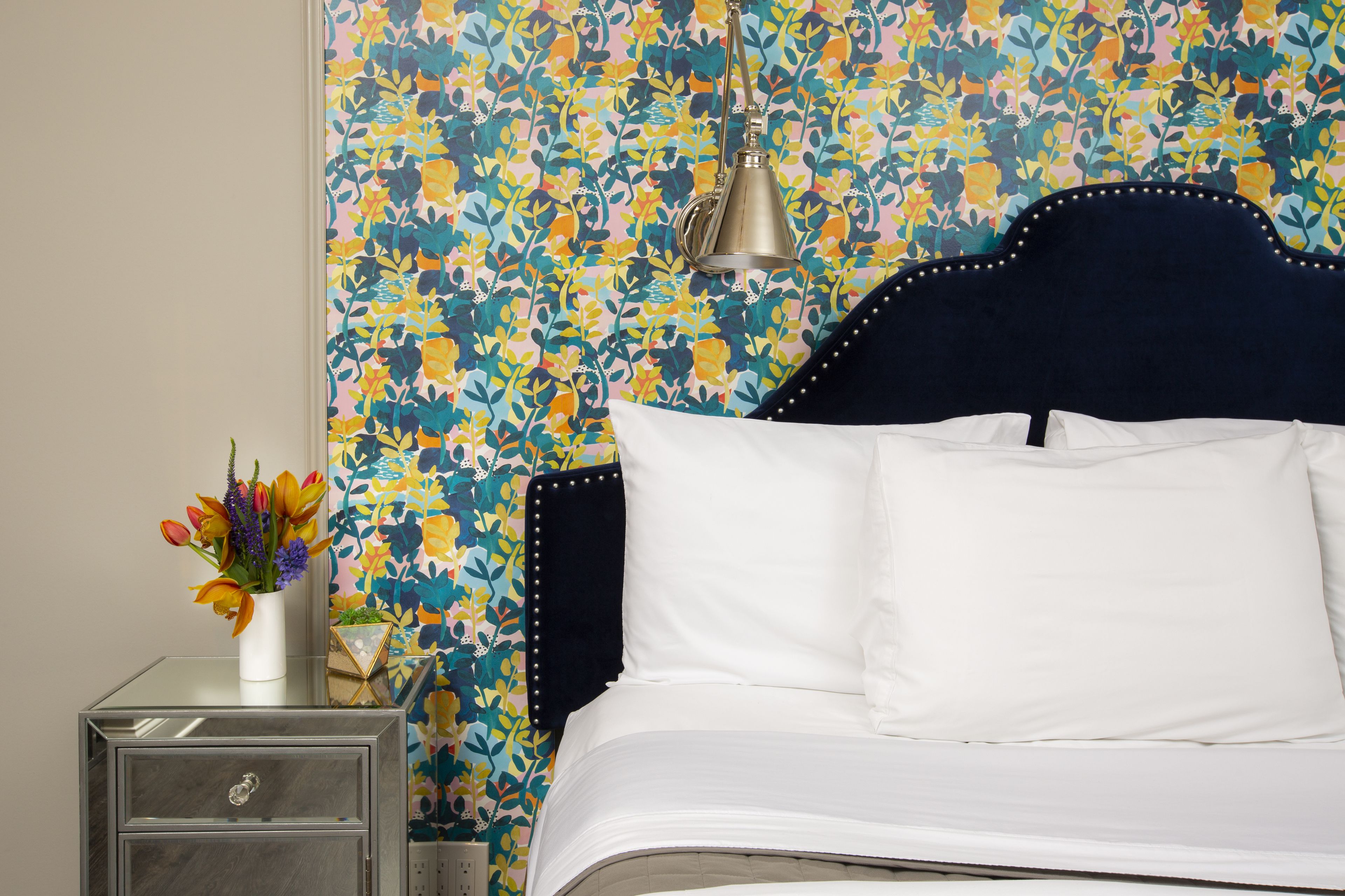 Una cama bien tendida con sábanas blancas, cabecera oscura y estampado de papel tapiz floral.