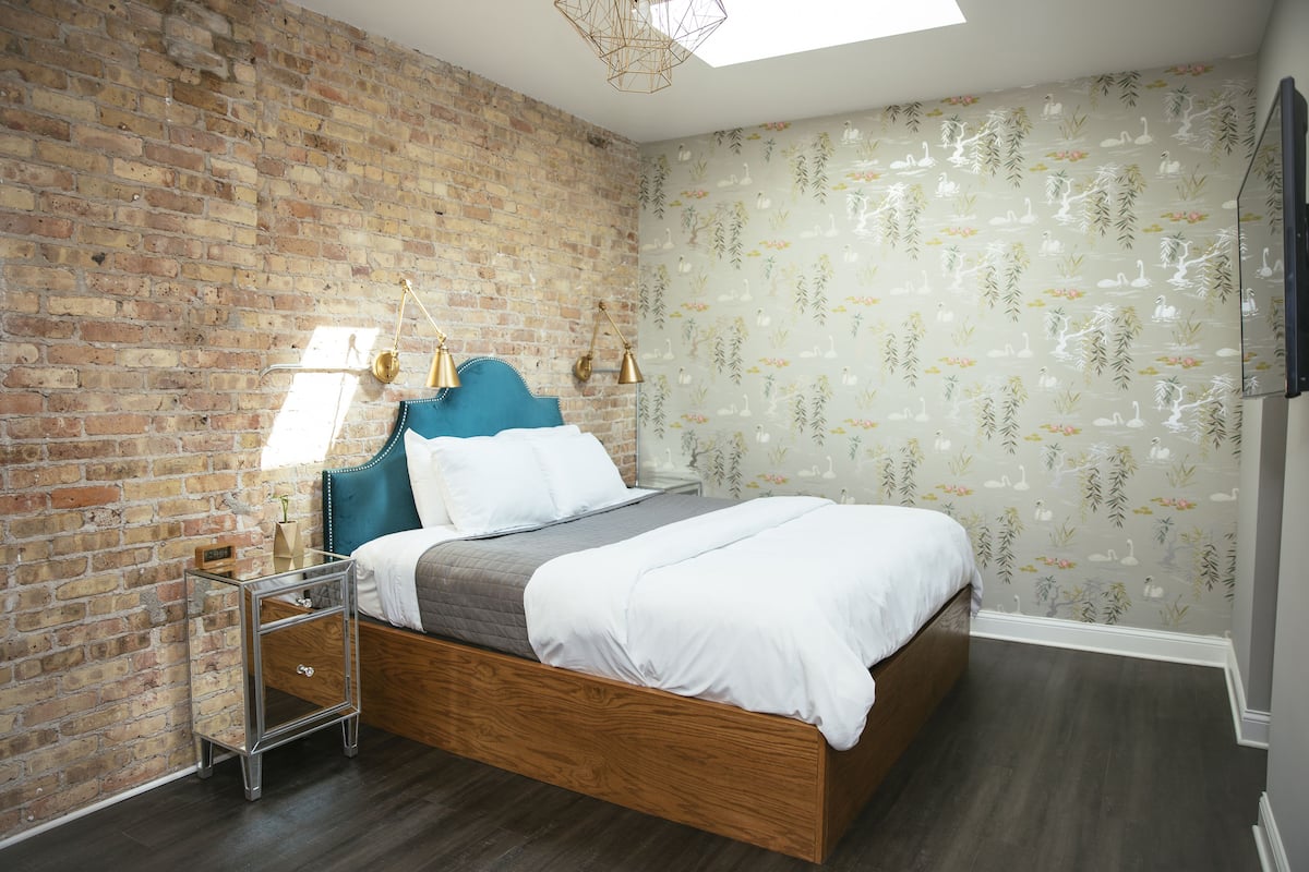 A bedroom with a brick wall, a bed with a blue headboard, a bedside table with lamps, and floral wallpaper.
