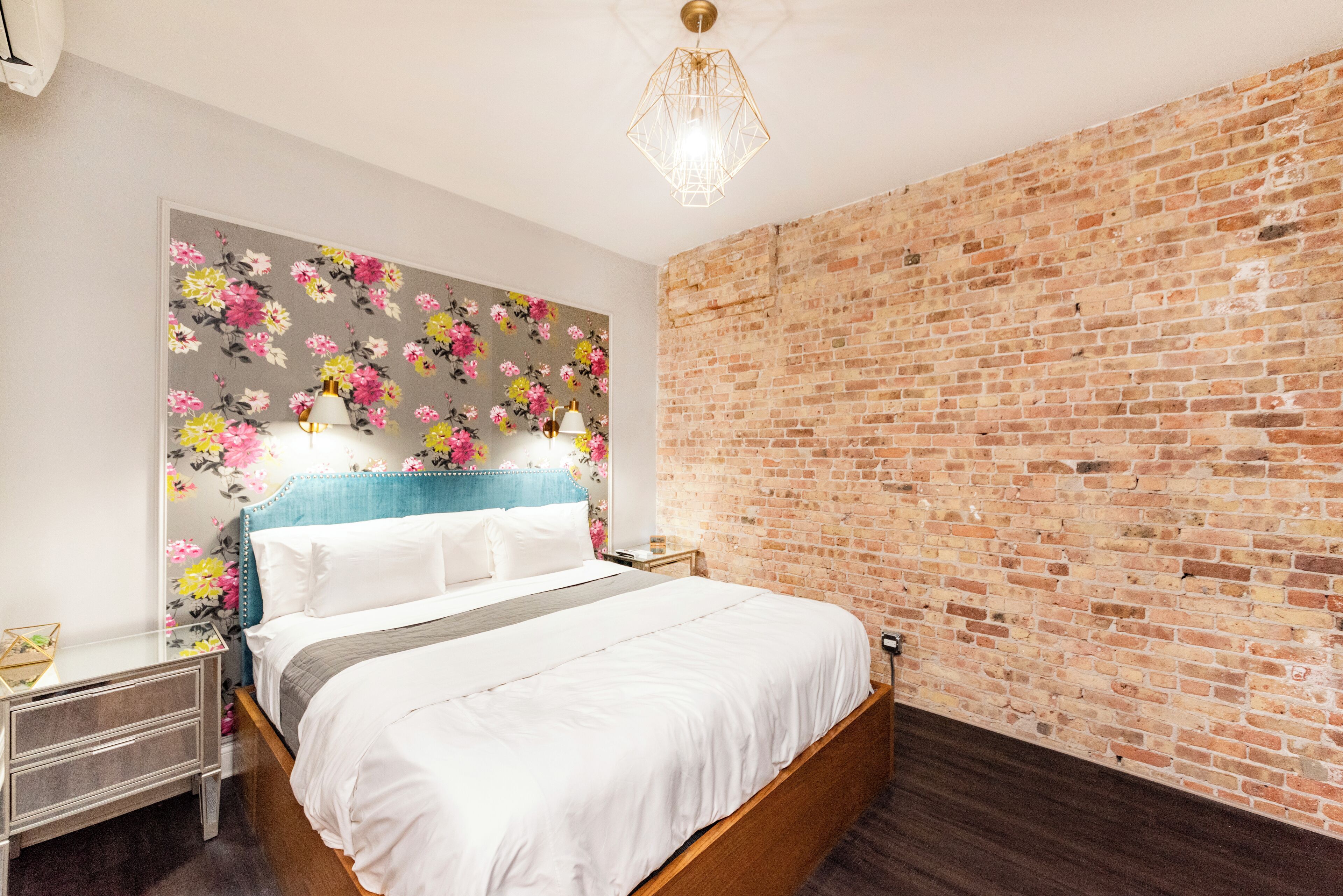 A bedroom with a brick wall, a floral headboard, a wooden bed frame, and a nightstand with a lamp.