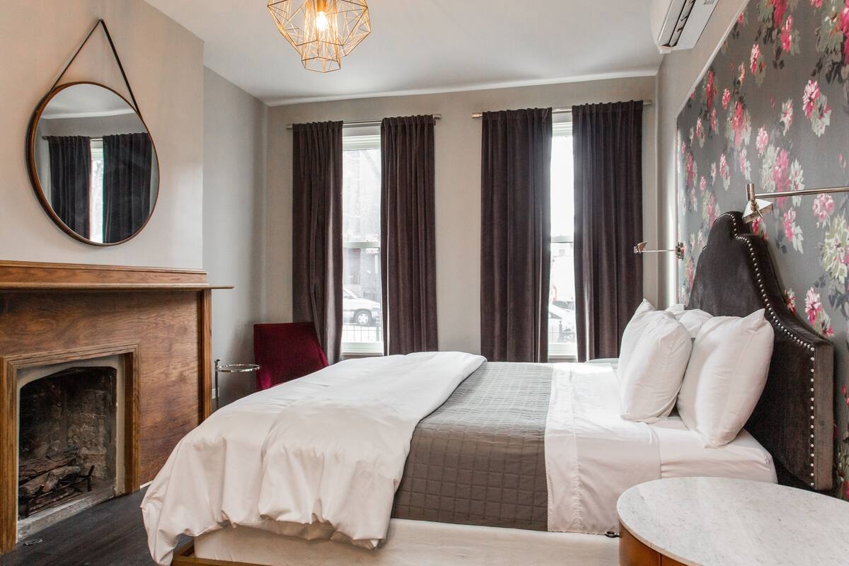 A bedroom with a large bed, a fireplace, and a view of the street through the window.