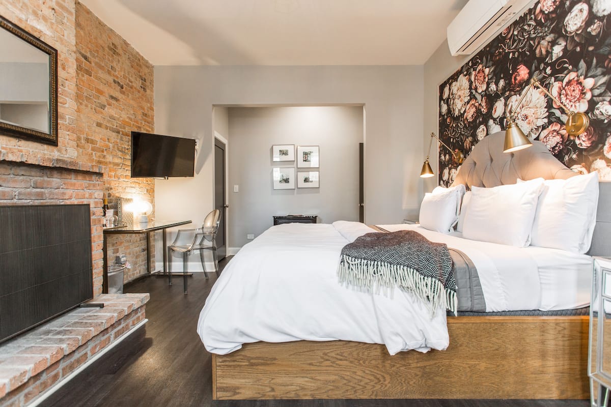 A bedroom with a brick wall, a flat-screen TV, a bed with white bedding, a floral wall tapestry, and a small table with chairs.