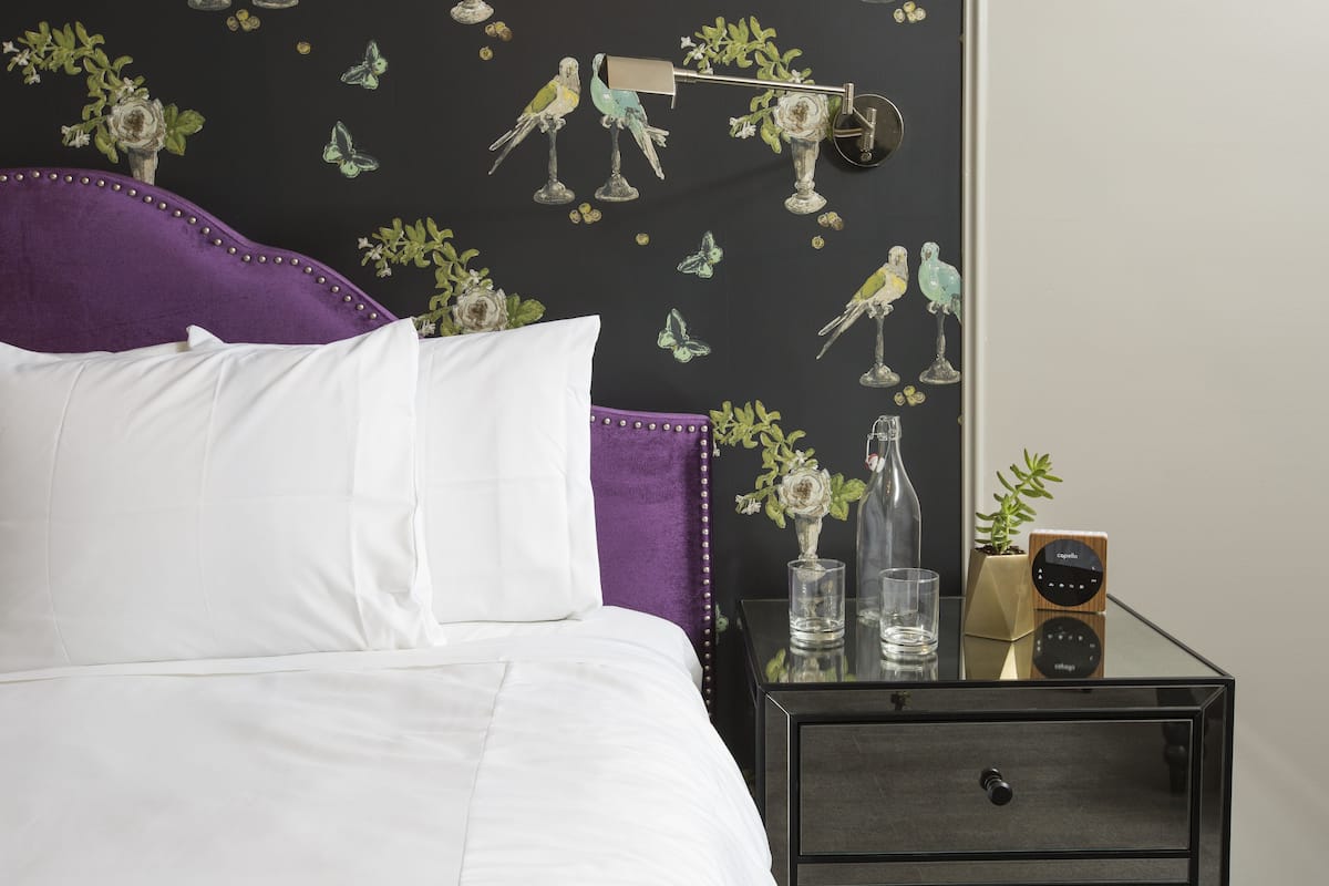 A neatly made bed with white linens and a purple headboard, a bedside table with a clock and a vase, and a wall with bird and flower decals.