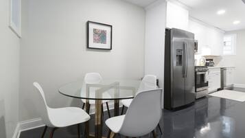 Une cuisine moderne équipée d’un réfrigérateur en acier inoxydable, d’un four encastré et d’un four à micro-ondes. On y trouve une table en verre et quatre chaises blanches.