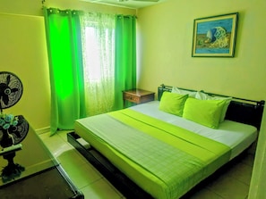 A hotel room with a bed, a nightstand, a fan, and a painting on the wall.