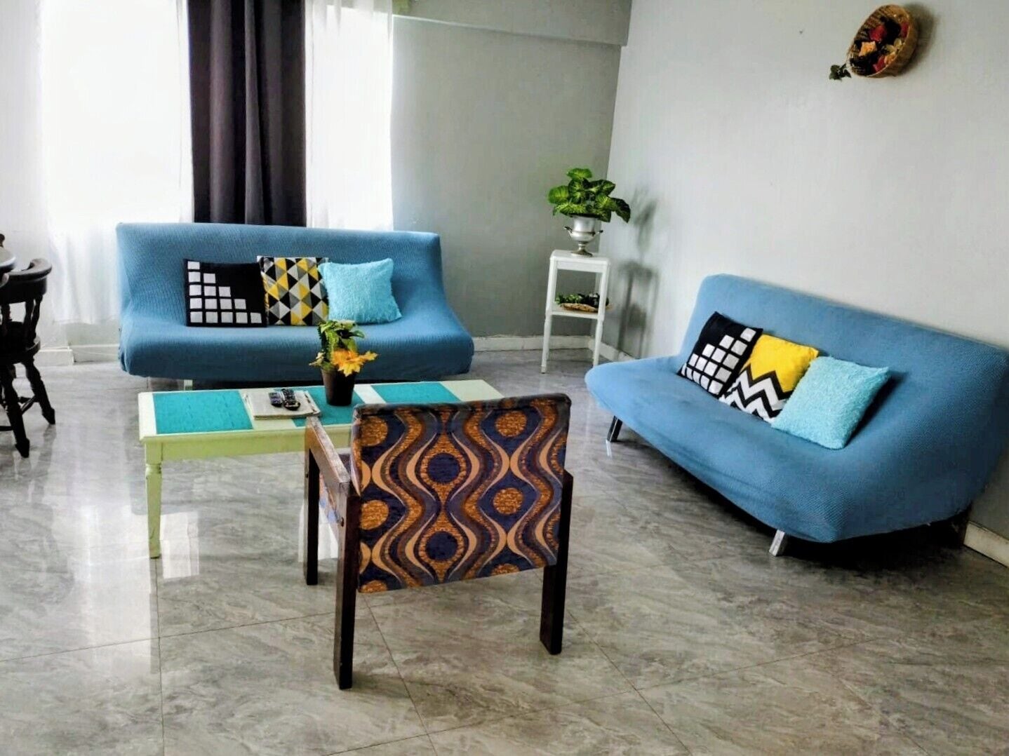 A living room with two blue sofas, patterned armchairs, a small table with a vase of flowers, and a potted plant on a white side table.