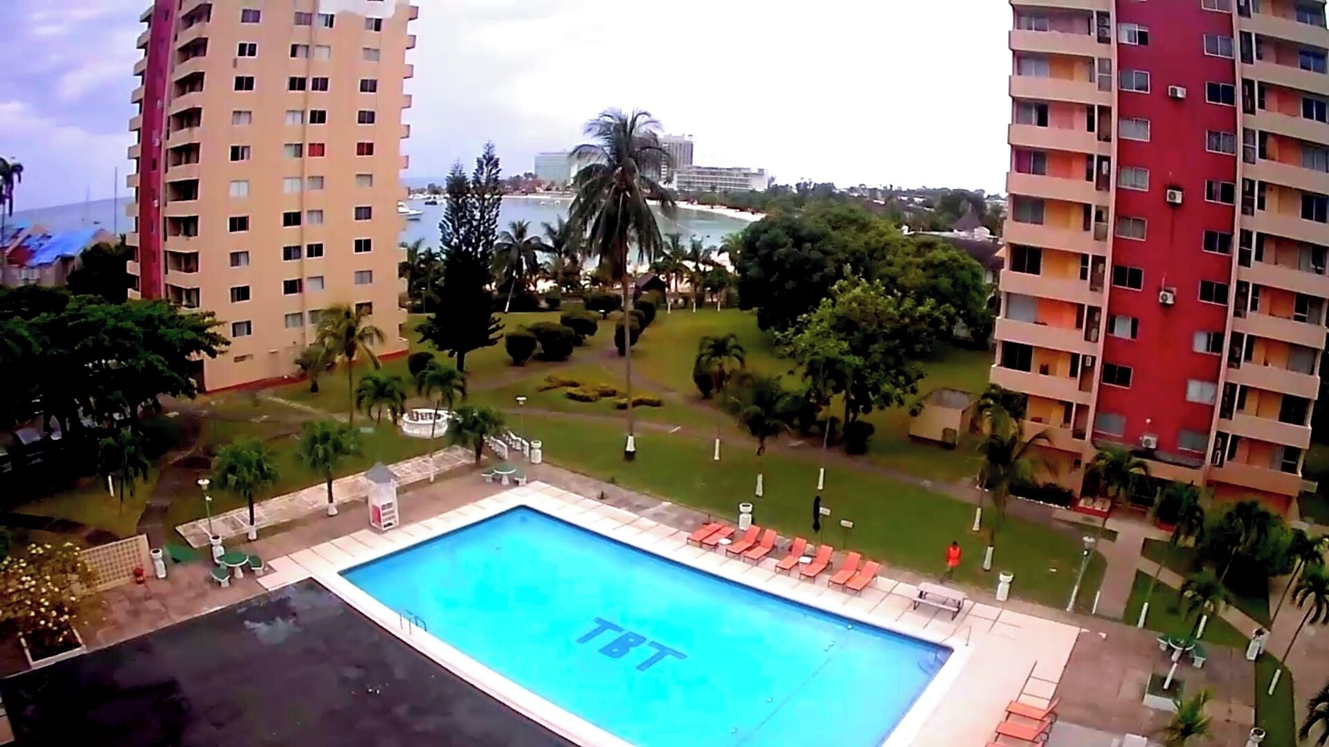 A swimming pool with 'TBT' text, surrounded by greenery and tall buildings.