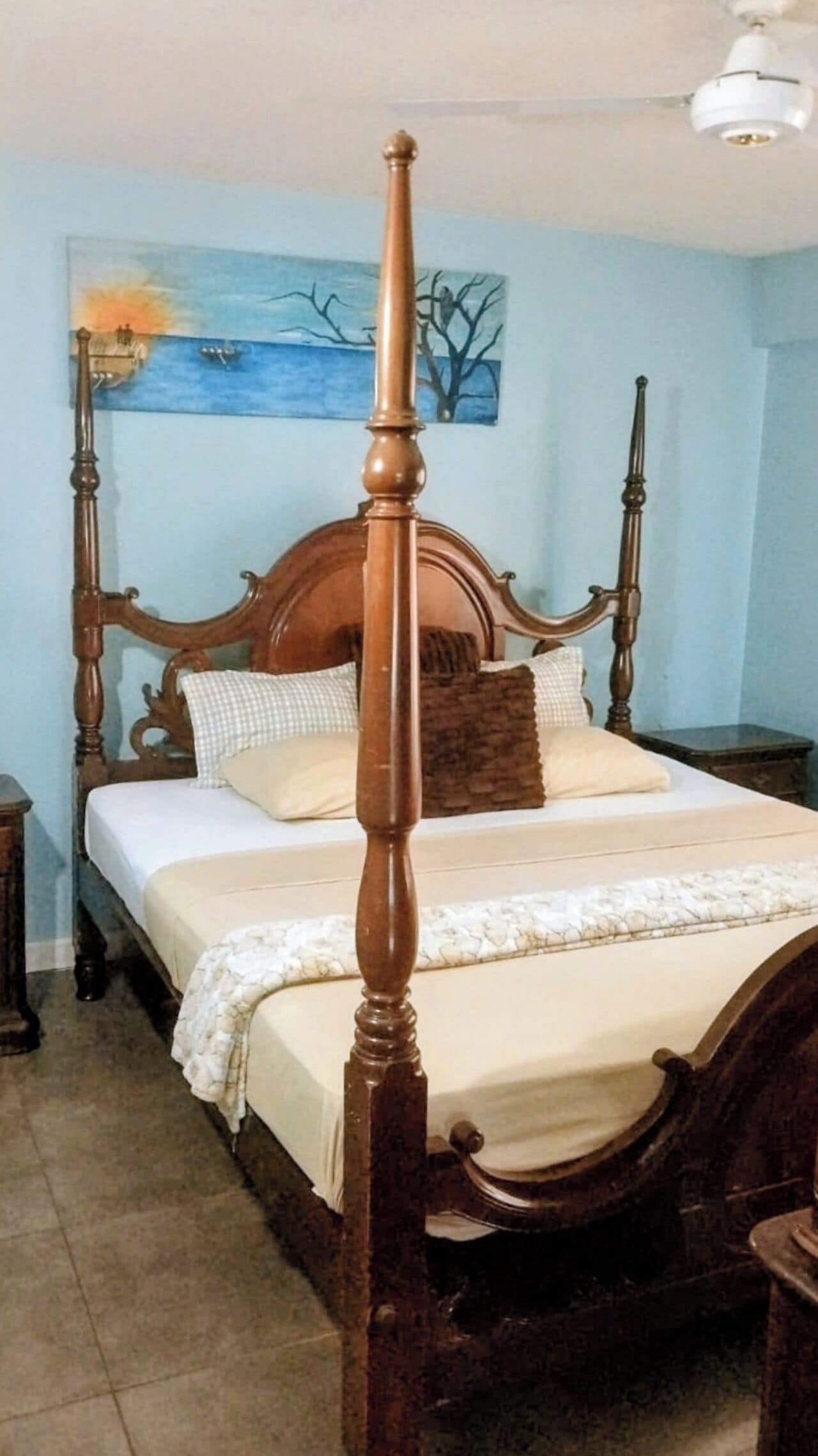 A four-poster bed with a wooden frame, a painting on the wall, and a ceiling fan.