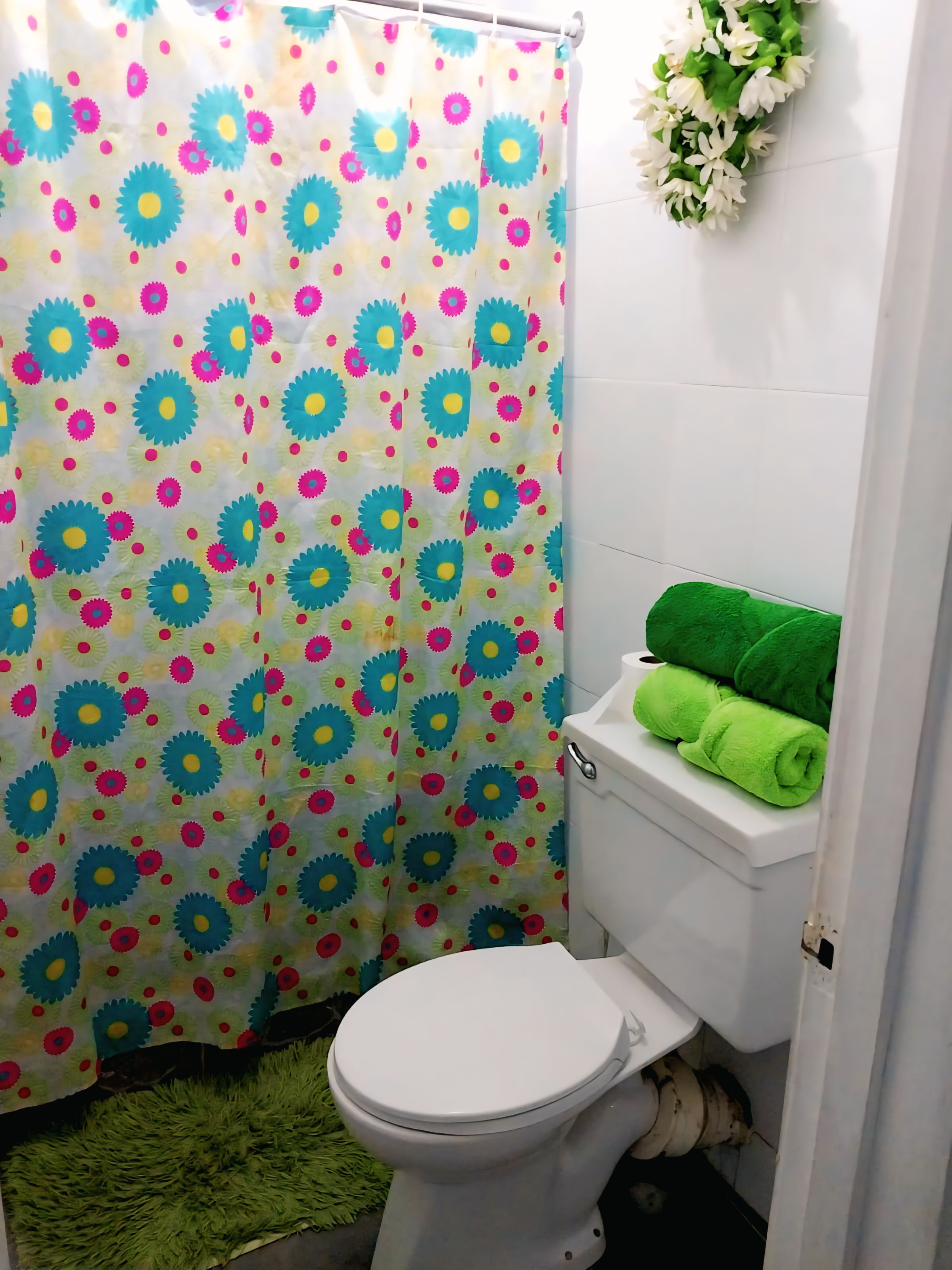 Casa de banho com sanita, toalha verde no tanque, cortina de chuveiro com padrão floral e tapete verde.