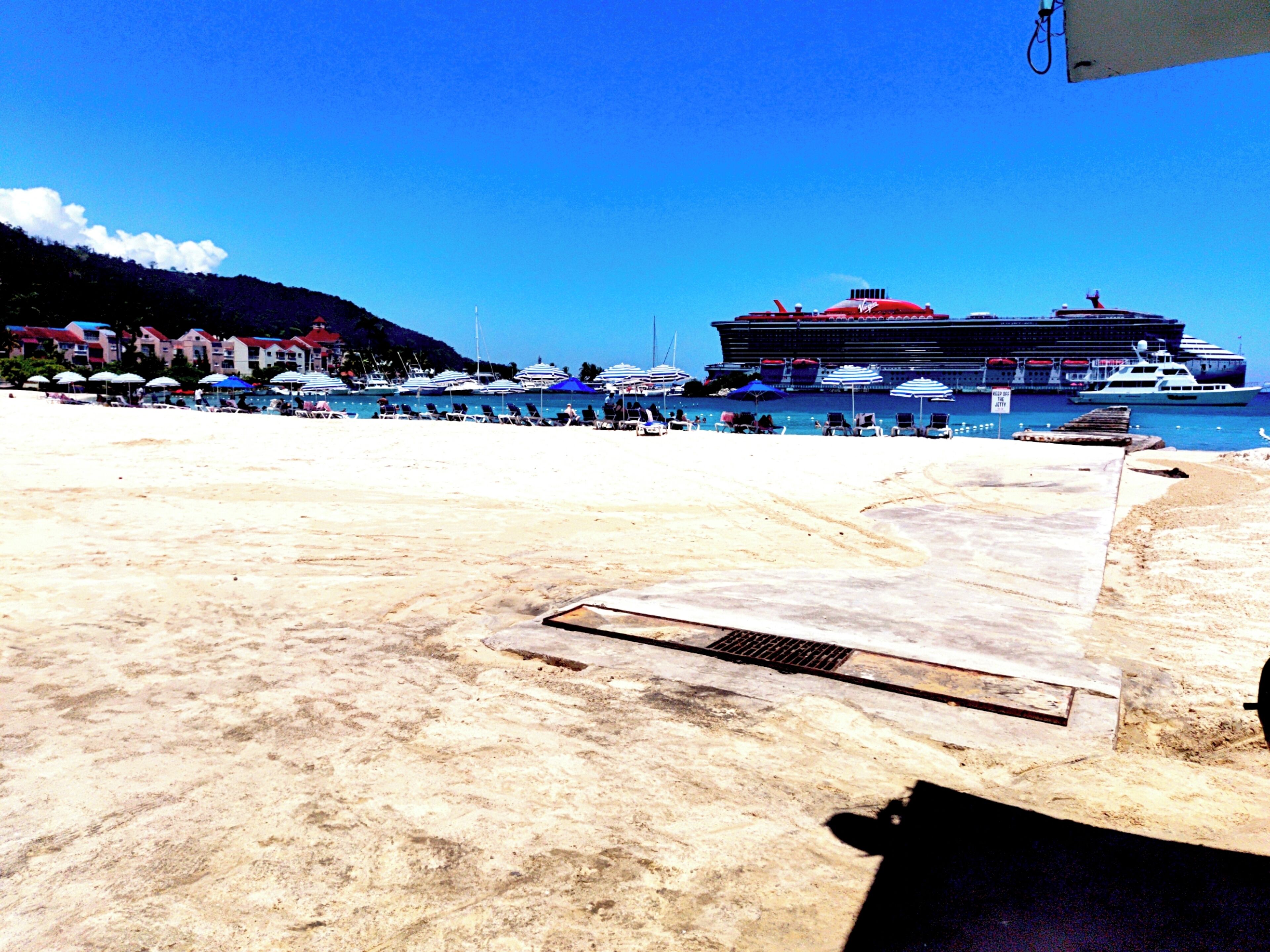 Ein Strand mit einem großen Kreuzfahrtschiff, Sonnenschirmen und einem sandigen Ufer.