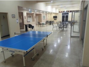 Une table de ping-pong se trouve dans une pièce où il y a des chaises, des tables, une porte et un téléviseur accroché au mur.