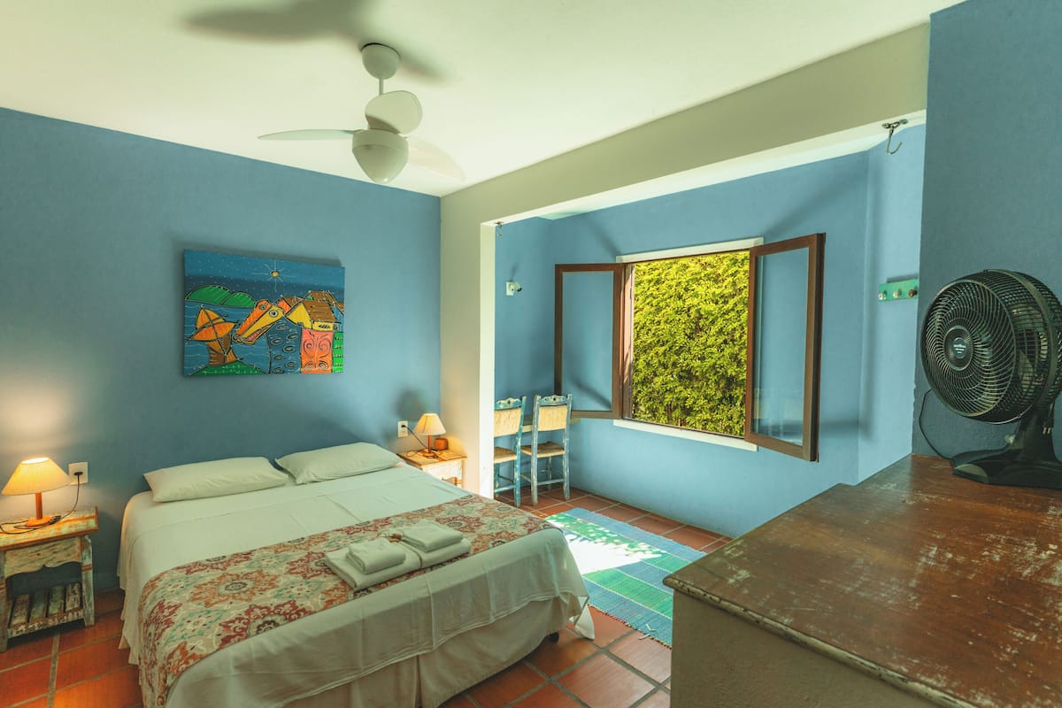 A bedroom with a bed, a ceiling fan, a window with a view of greenery, a desk with a fan, and a painting on the wall.