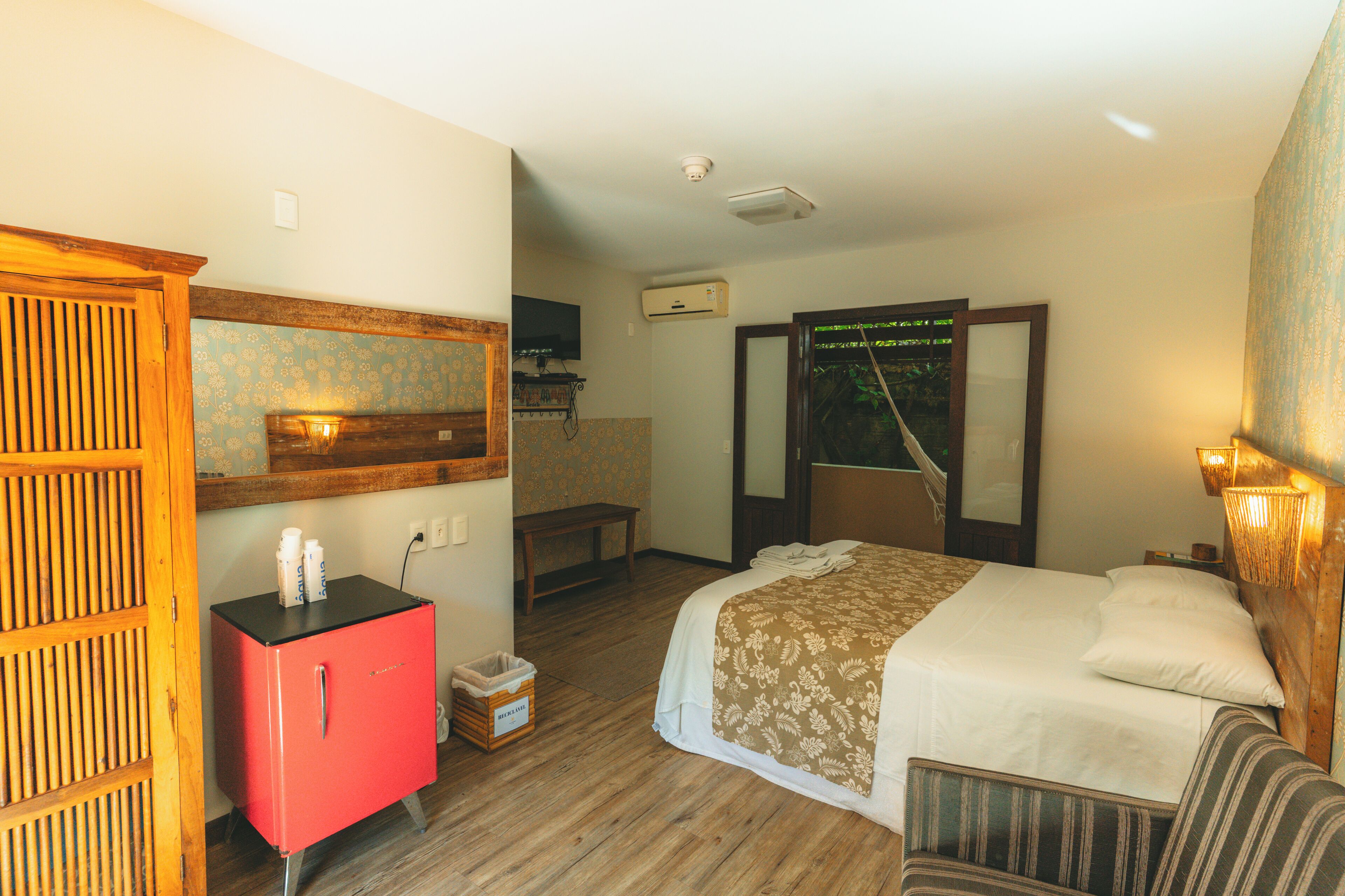 Une chambre d’hôtel avec un lit, un mini-bar rouge, une armoire en bois, un banc et un miroir.