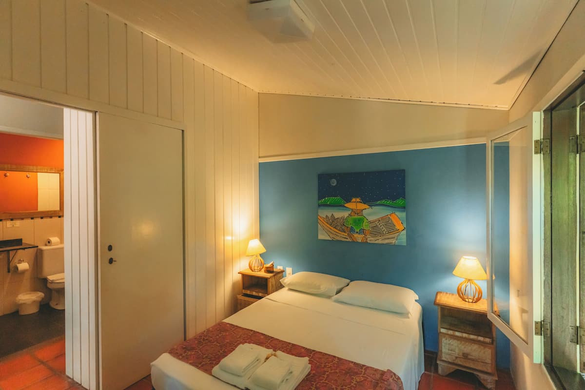 A hotel room with a bed, two bedside tables with lamps, a painting on the wall, and a bathroom visible through an open door.