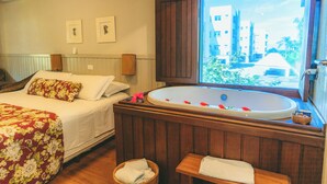 Un dormitorio con una bañera grande, una cama con ropa de cama floral y una ventana con vistas a edificios y vegetación.
