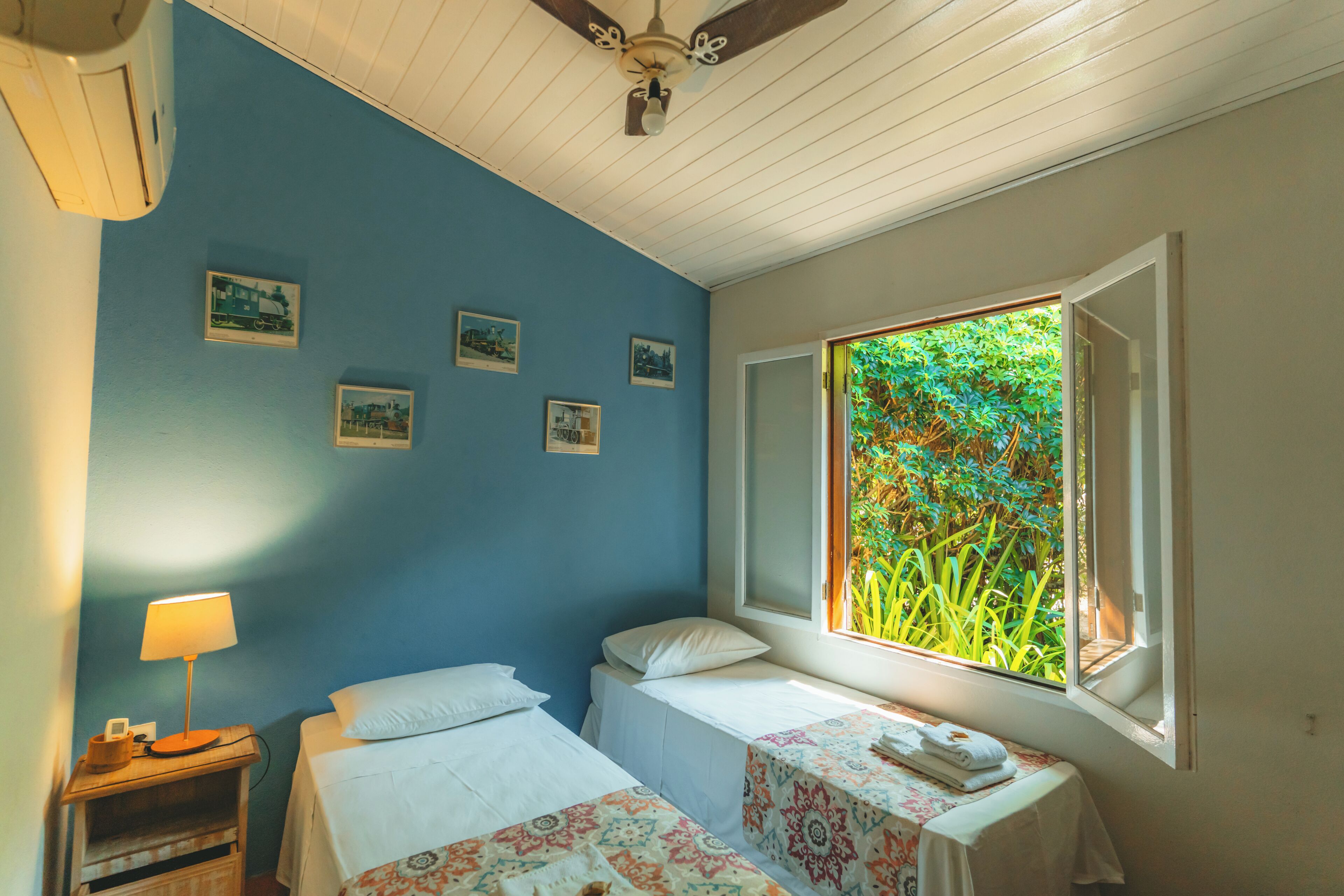 A room with two beds, a window with a view of greenery, a ceiling fan, and a wall with framed pictures.