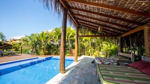 Un área de piscina con zona de estar techada, piscina y palmeras circundantes.