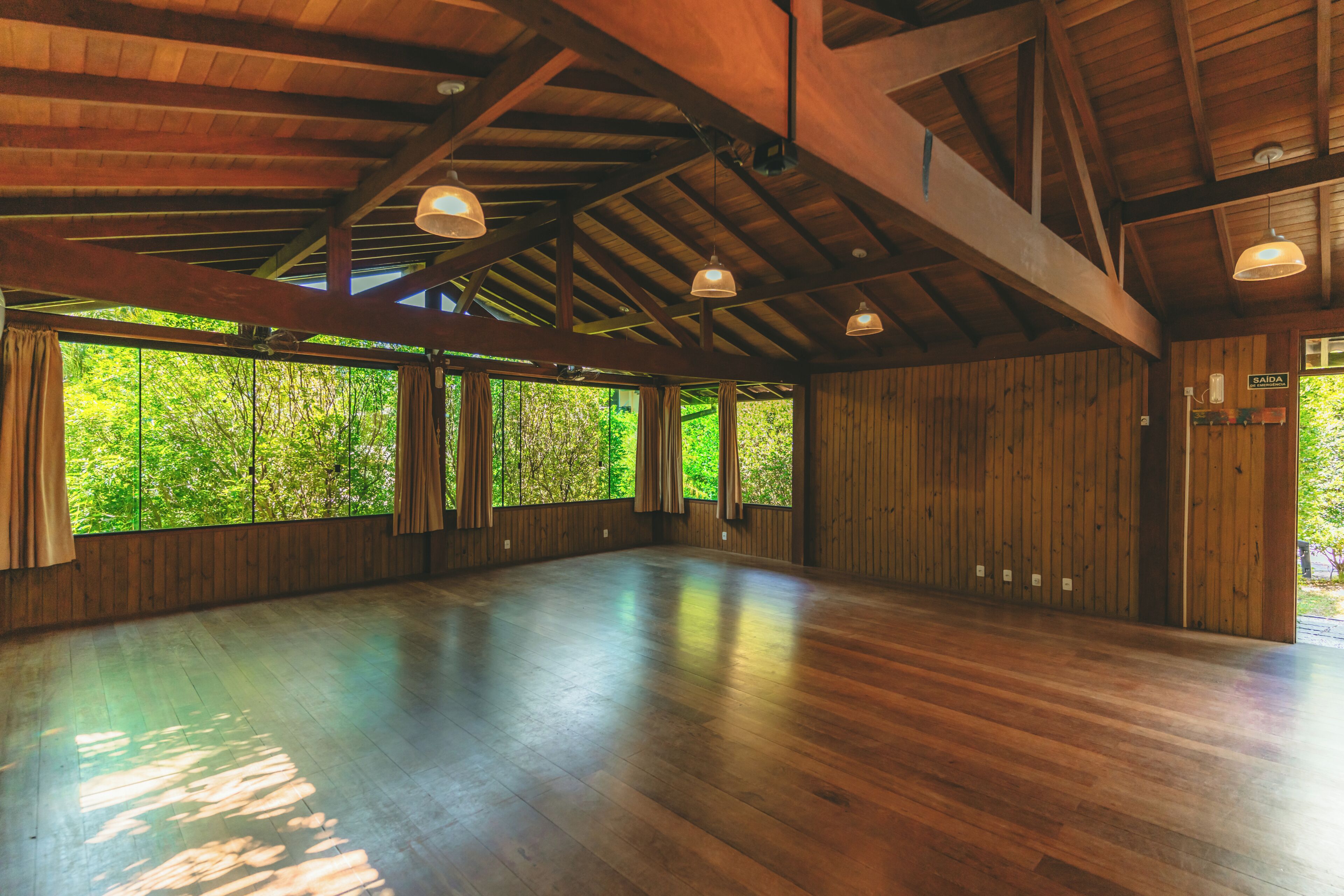 A spacious room with wooden floors and ceiling beams, large windows with curtains, and hanging pendant lights.