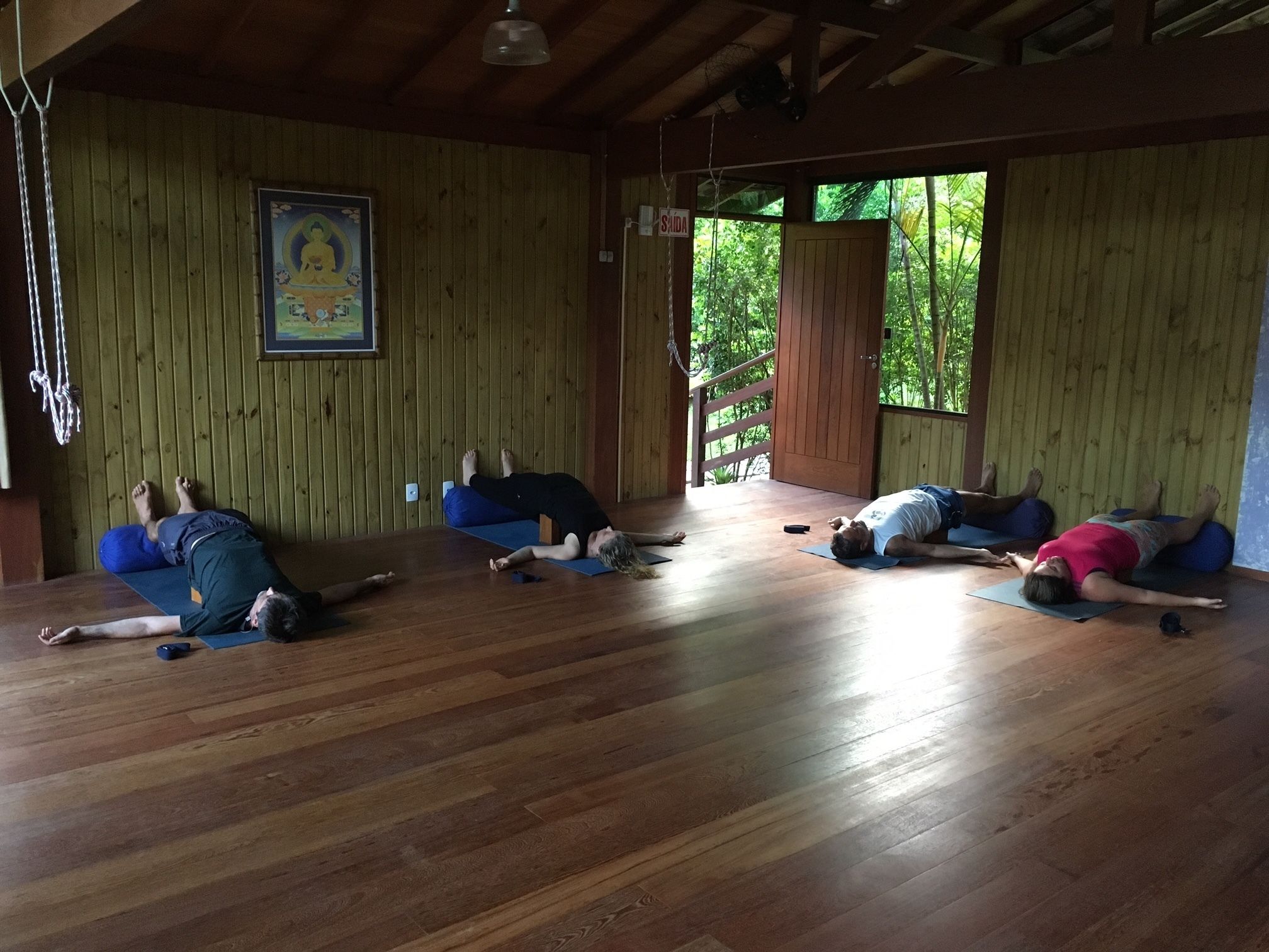 Um estúdio de yoga com paredes e chão de madeira. Quatro pessoas praticam yoga em tapetes.