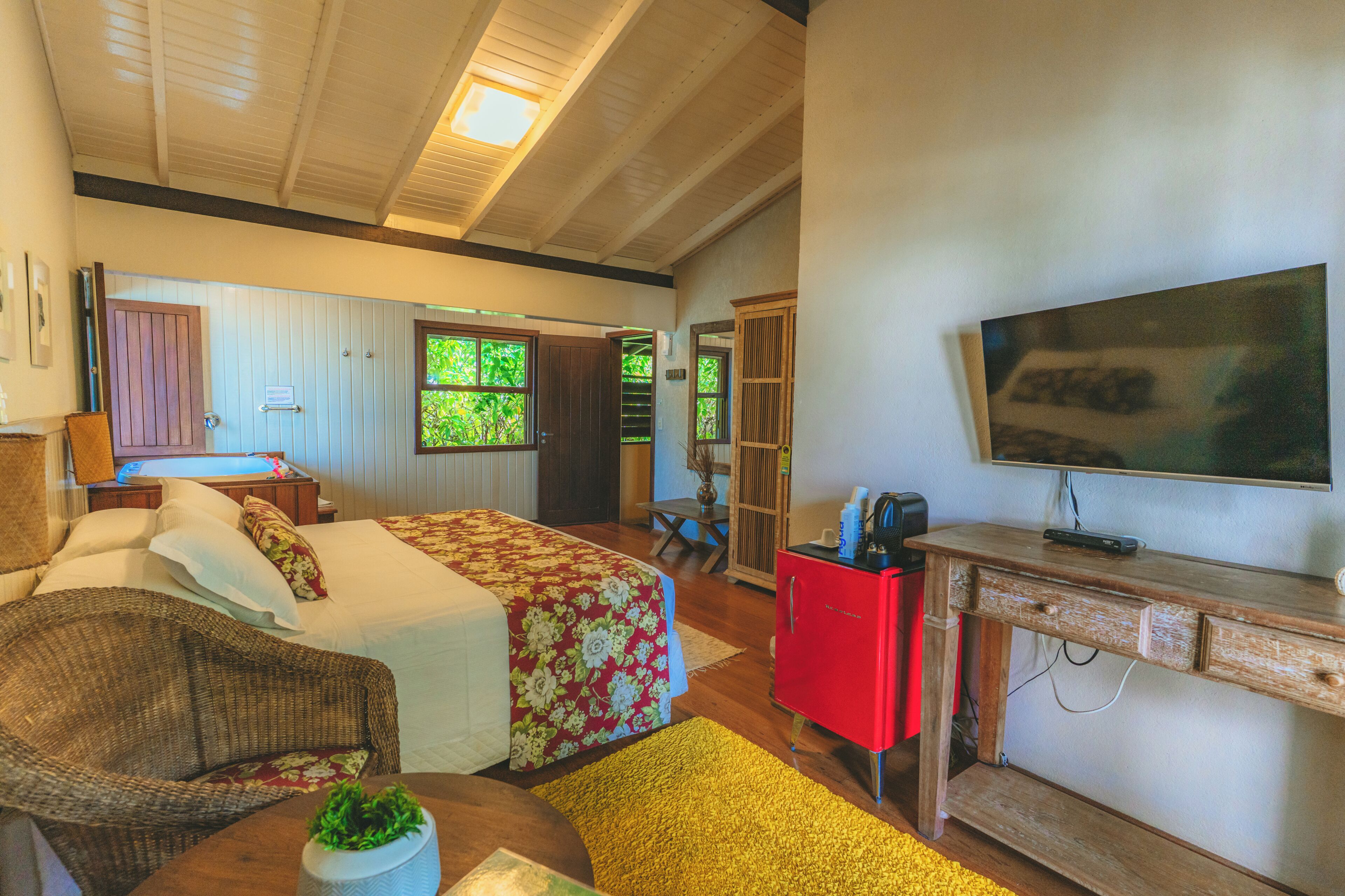Une chambre d’hôtel avec un lit, un bureau avec une télévision, une chaise en osier et une petite table avec une plante.