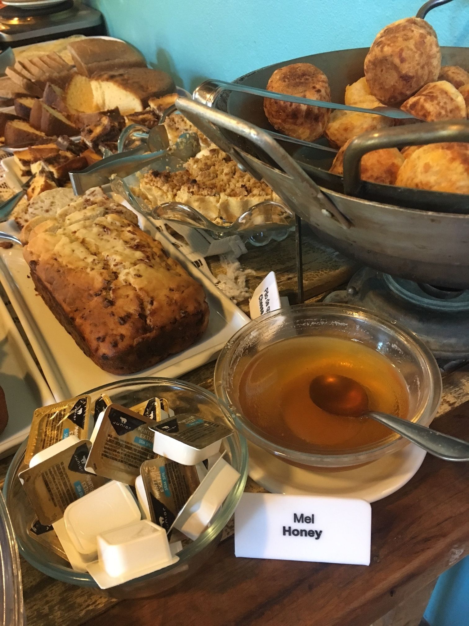 A buffet with various baked goods, including bread and muffins, and a bowl of honey labeled "Mel Honey".
