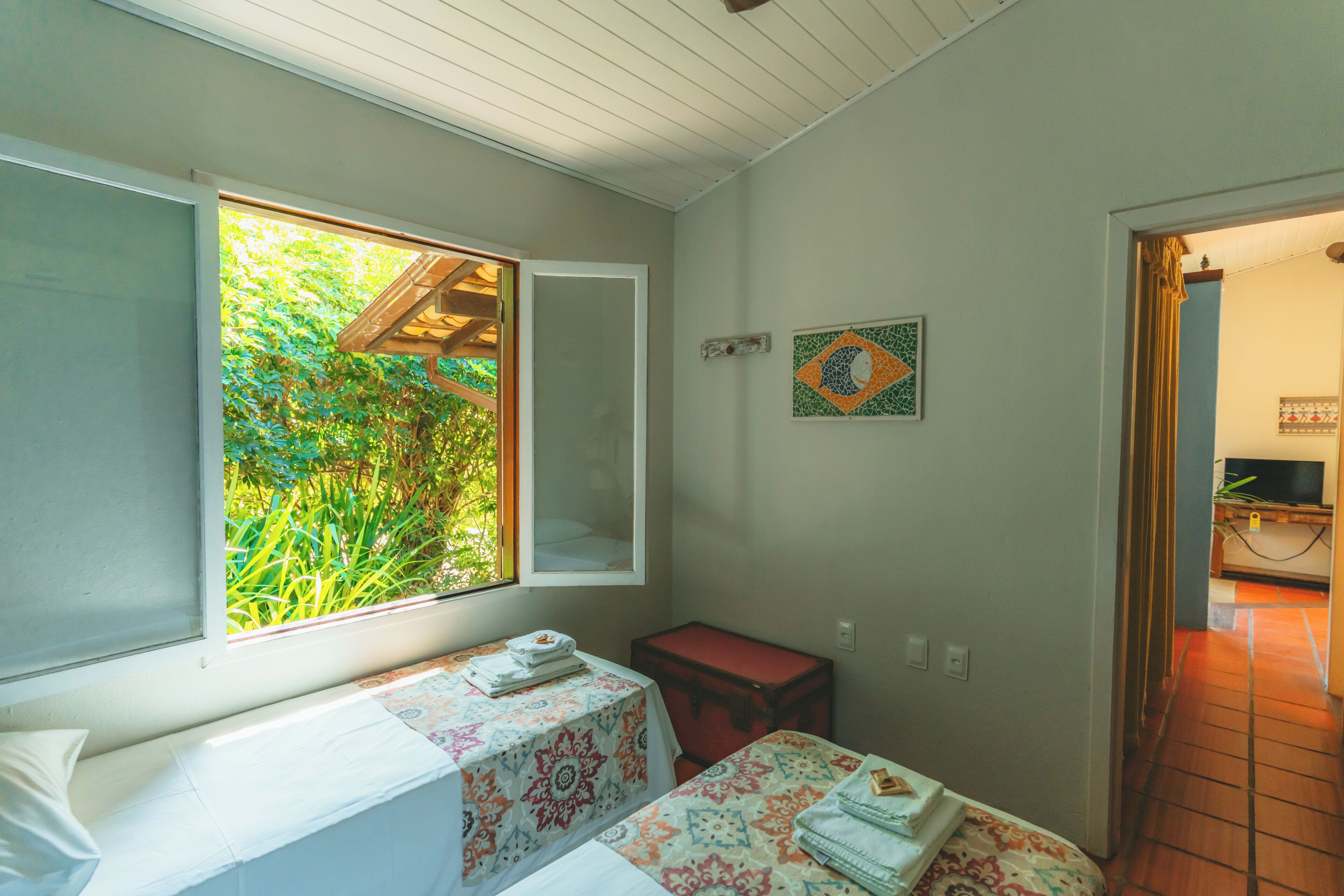 A room with two beds, a window with a view of greenery, a small table, and a red suitcase.