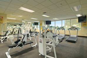 Une salle de sport équipée de divers appareils d’exercice, avec un téléviseur fixé au mur et de grandes fenêtres donnant sur l’extérieur.
