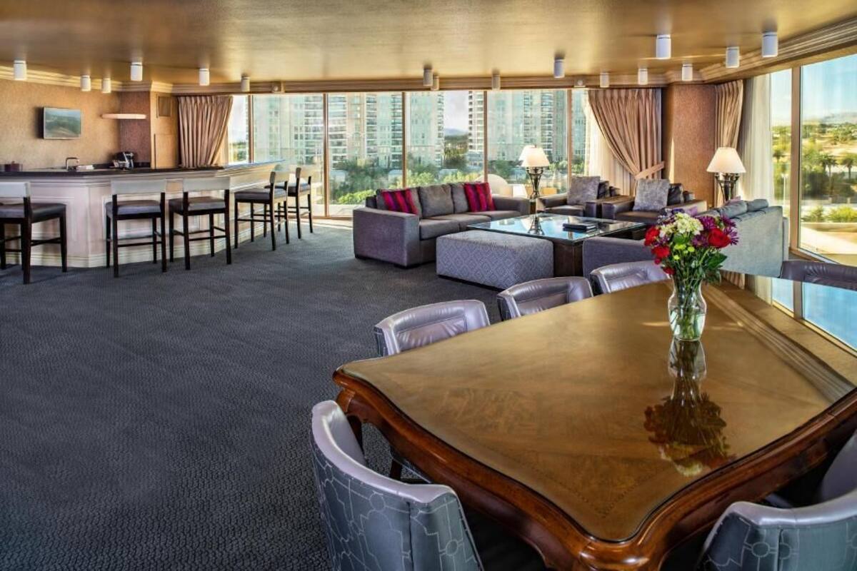 Amplia habitación de hotel con zona de comedor, un conjunto de sofás y un gran ventanal con vistas a la ciudad.