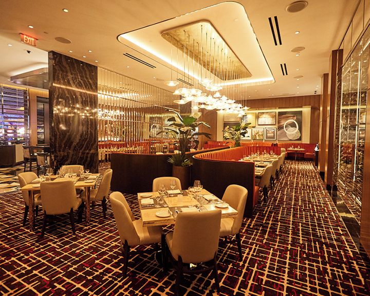 Área de jantar de hotel com mesas preparadas para hóspedes, carpete estampado e um lustre moderno.