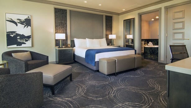Habitación de hotel con una cama grande, dos mesitas de noche con lámparas, una zona de estar con sofá y sillón, y un baño visible a través de una puerta abierta.