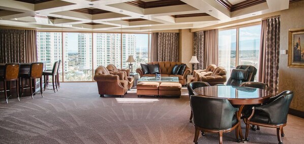 A spacious hotel room with a large seating area, a dining table, and a view of the cityscape through large windows.