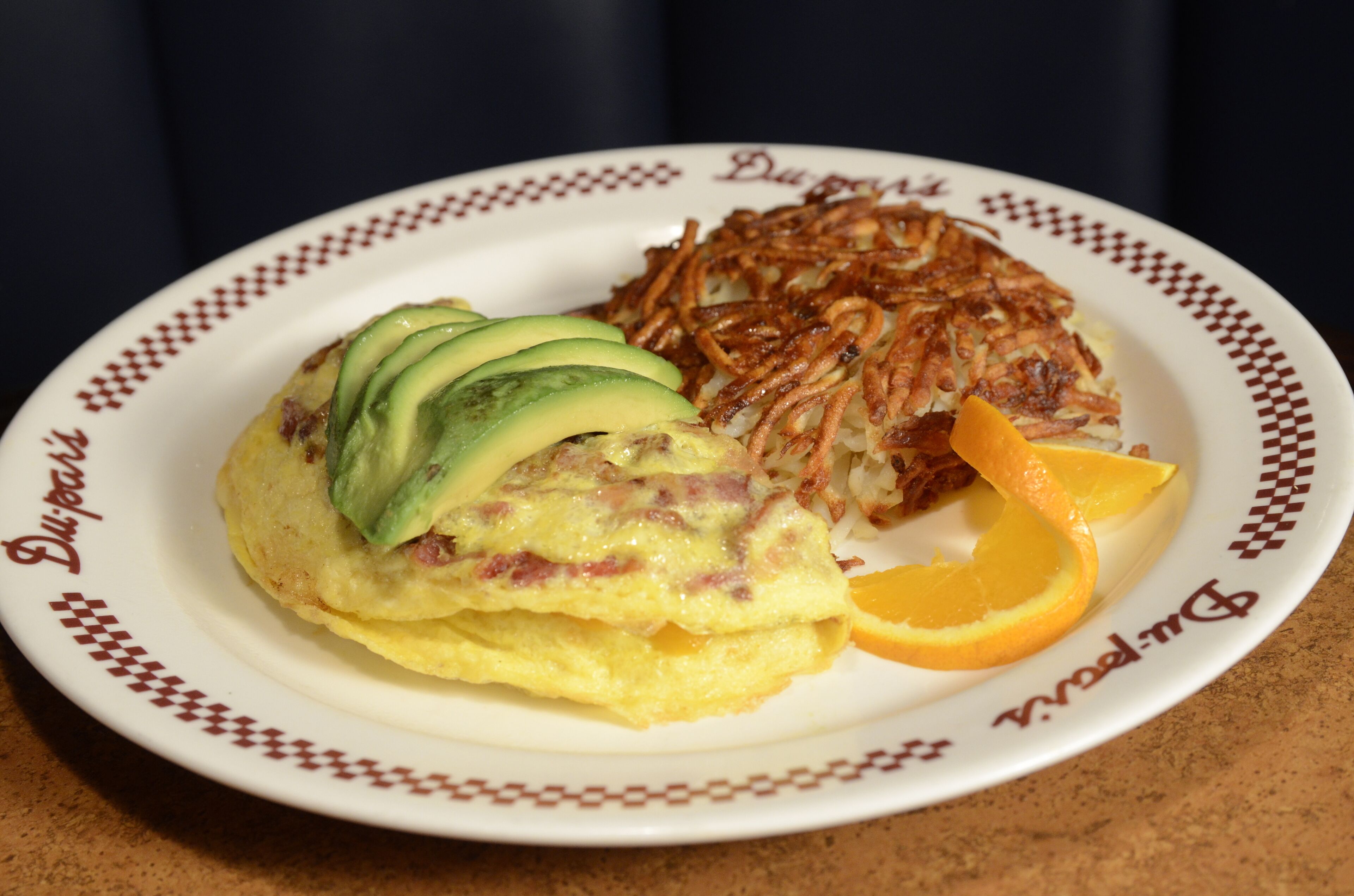 Une assiette contenant une omelette, des pommes de terre rissolées, des tranches d’avocat et une tranche d’orange.