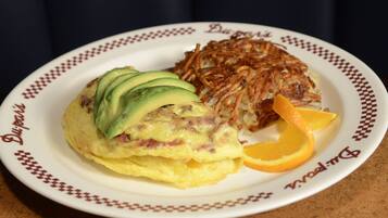 Une assiette contenant une omelette, des pommes de terre rissolées, des tranches d’avocat et une tranche d’orange.