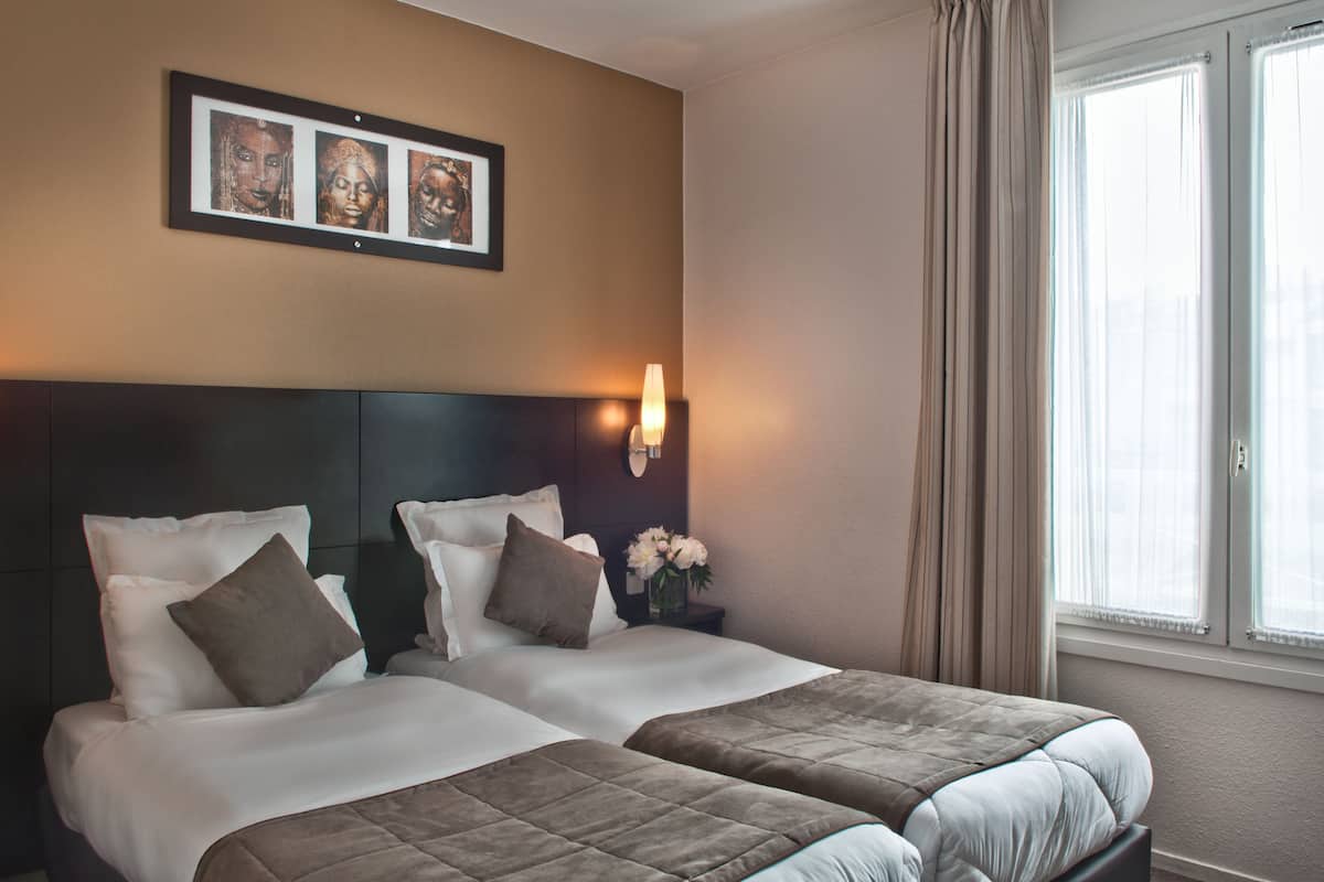 Habitación de hotel con dos camas, cabecero oscuro, lámpara de pared y un cuadro enmarcado sobre las camas, además de una ventana con cortinas.