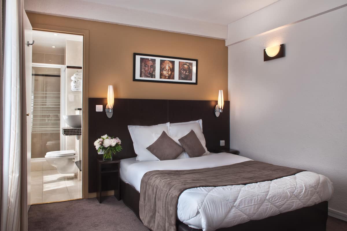 Una habitación de hotel ordenada con una cama grande, una mesita de noche con un florero, una obra de arte enmarcada sobre la cama y un vestidor con ropa colgada.