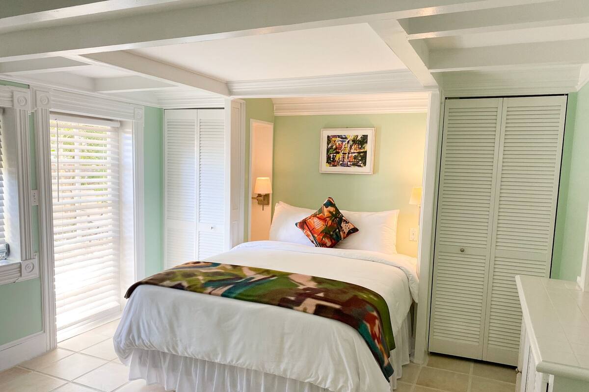 A bedroom with a bed, a window with blinds, a door, and a wall with a picture.