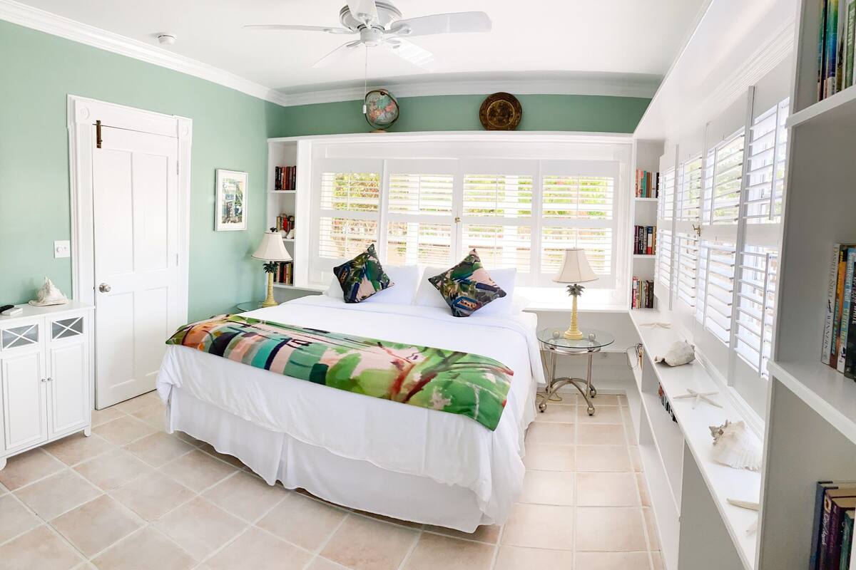 A bedroom with a bed, a window with blinds, a bedside table, a lamp, and a bookshelf.