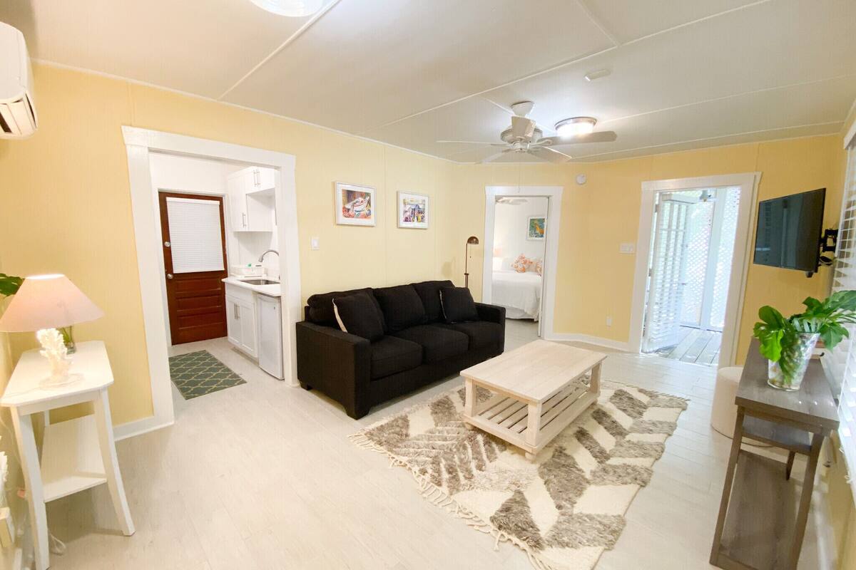 A living room with a black sofa, a wooden coffee table, a small white table with a lamp, a kitchen area, and a door leading to a bedroom.